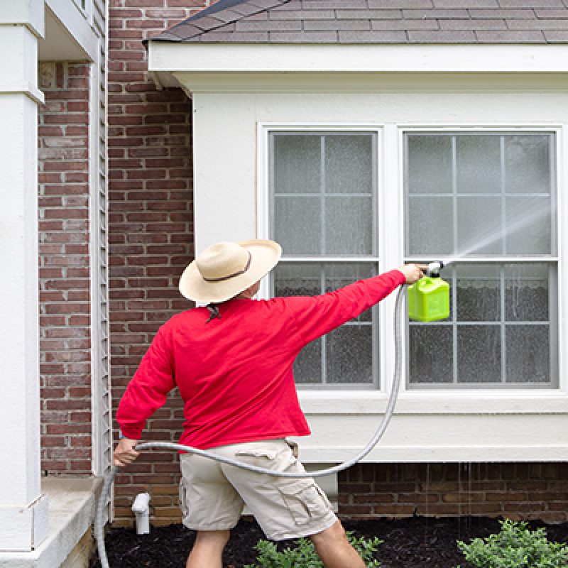 Man spring cleaning the exterior of his house washing down a bay window with a high pressure sprayer attached to a hose pipe in a close up rear view